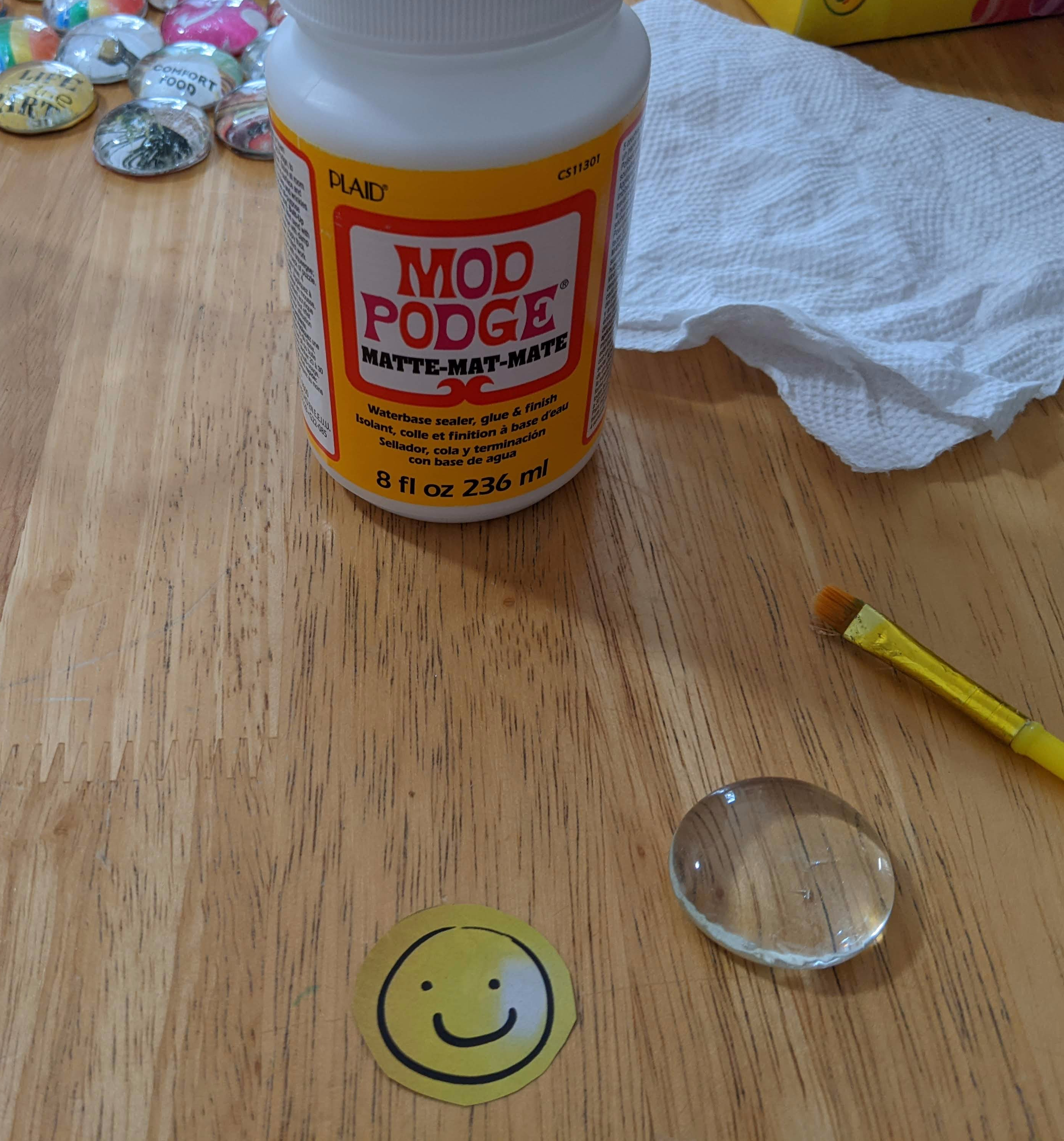 photo of bottle of Mod Podge next to a paper towel, a small yellow paintbrush, a glass gem, and a photo of a smiley face